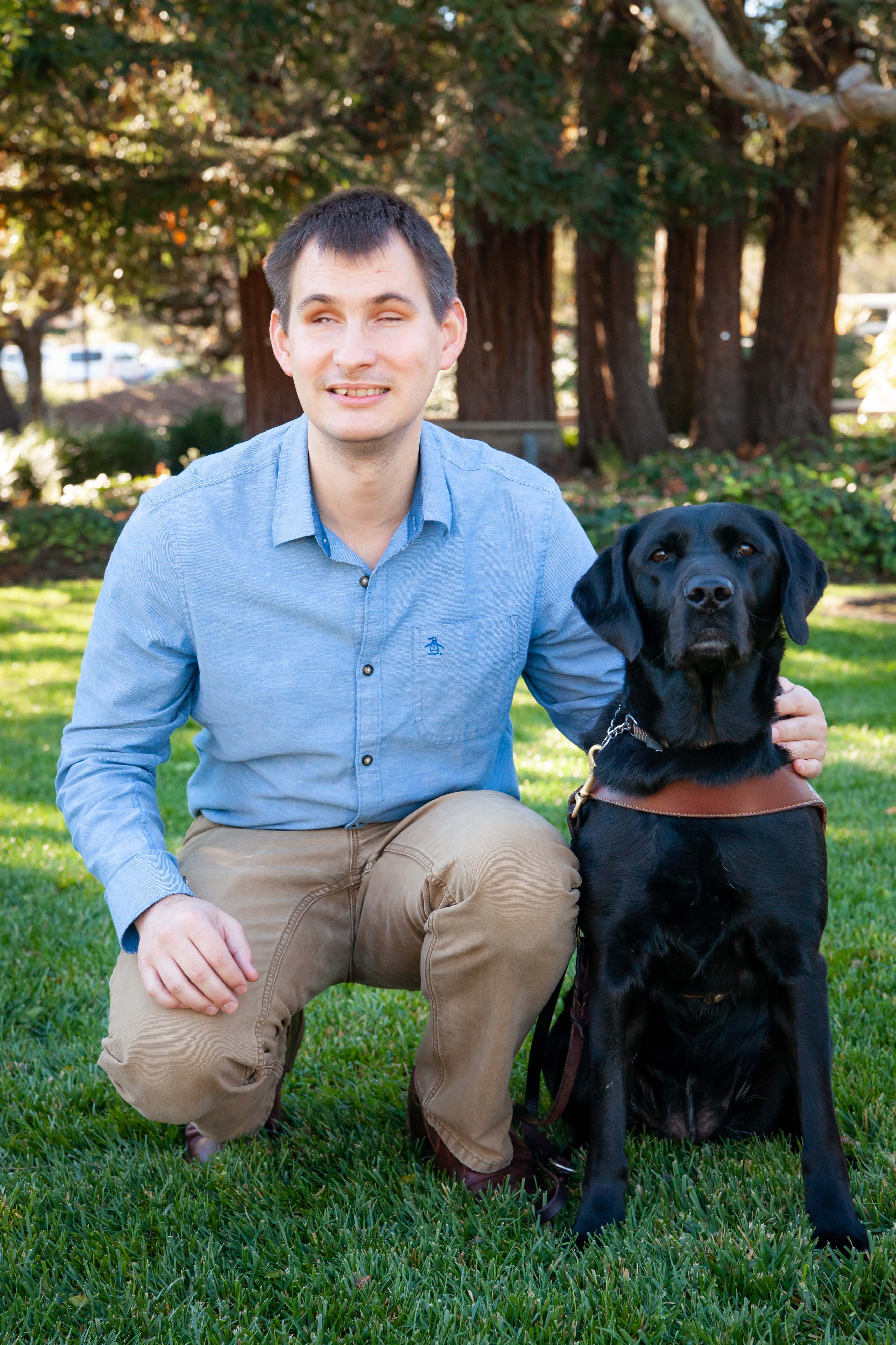 Jake Koch from Guide Dogs for the Blind and his guide dog, Forli. Jake and Forli are standing in a grassy area with trees in the background. Jake is wearing a blue button down shirt and brown pants and is squatting next to Forli, who is a black labrador retriever.
